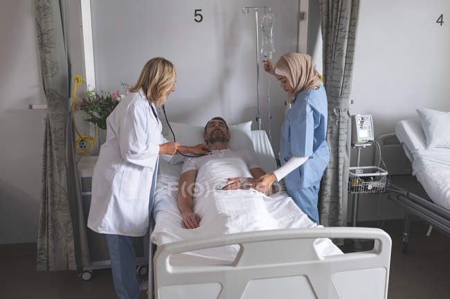Vista lateral del médico femenino caucásico que examina al paciente varón caucásico con estetoscopio en la sala del hospital. En el lado derecho de la cama de raza mixta enfermera en hiyab está ayudando a la doctora caucásica . - foto de stock