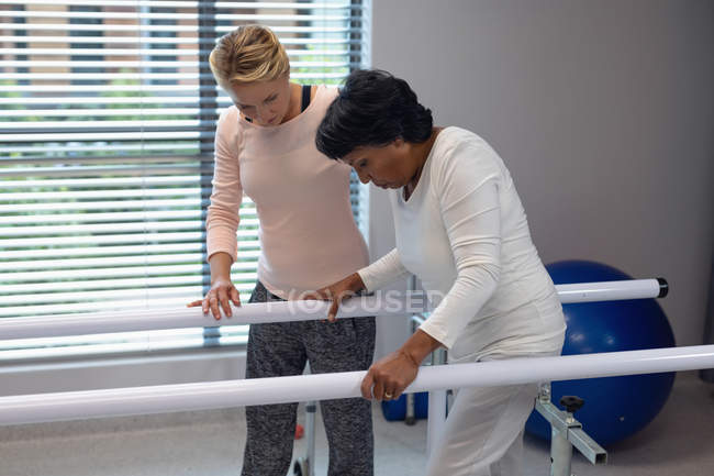Vista lateral del fisioterapeuta femenino caucásico ayudando a la paciente de raza mixta a caminar con barras paralelas en el hospital - foto de stock