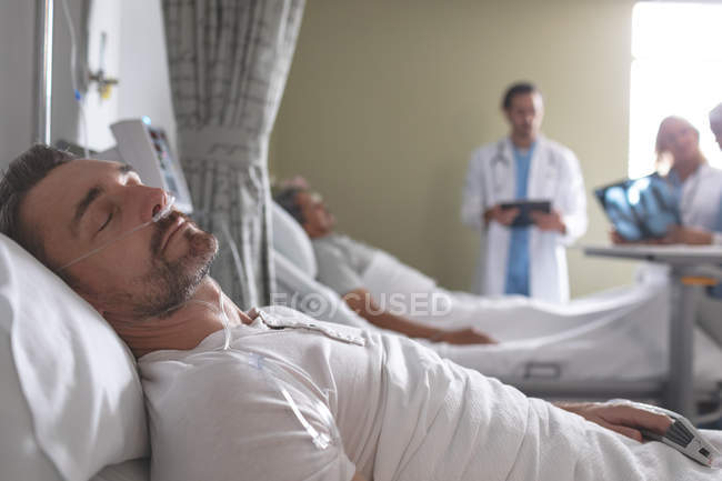 Vue latérale du patient de sexe masculin de race blanche qui dort sur son lit à l'hôpital. En arrière-plan, divers médecins discutent de rayons X avec un patient masculin métis . — Photo de stock