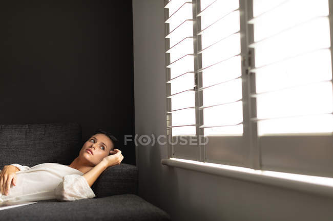 Боковий вид вдумливого кавказької жінки, що розслабляє на дивані у вітальні будинку — стокове фото