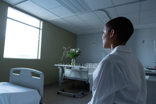Вид з боку вдумливого змішаних раси жіночого пацієнта сидять на ліжку в приході, дивлячись за вікном в лікарні. — стокове фото