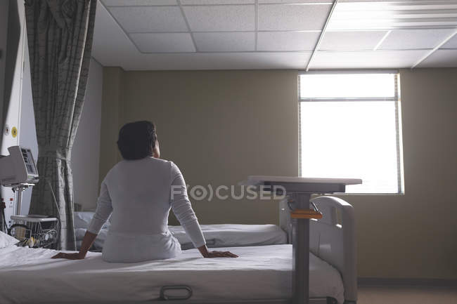 Rear view of mixed-race female patient sitting on bed while looking outside in the ward at hospital — Stock Photo