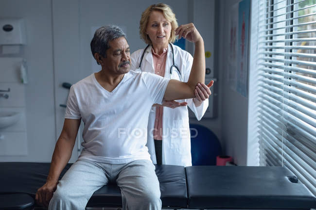 Vista frontal de la doctora caucásica examinando el brazo de paciente masculino de raza mixta senior en el hospital - foto de stock