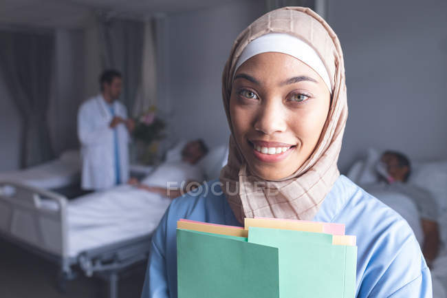 Porträt einer gemischten Ärztin im Hijab, die mit Krankenakten auf der Station im Krankenhaus steht. Im Hintergrund spricht kaukasischer Arzt mit Patient. — Stockfoto