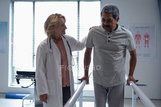 Vista frontal del médico caucásico ayudando a pacientes de raza mixta a caminar con barras paralelas en el hospital - foto de stock