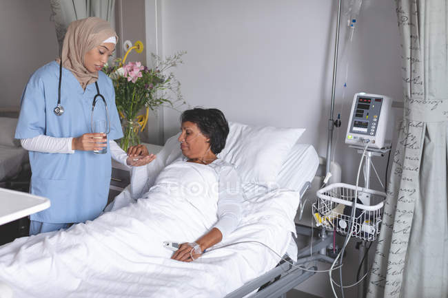 Вид спереду красивого лікаря змішаної раси в хіджабі, який дає ліки пацієнтці змішаної раси в лікарні . — стокове фото