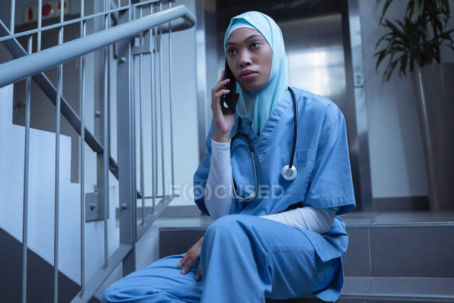 Обзор медсестры смешанной расы в хиджабе, разговаривающей по мобильному телефону, сидя на лестнице в больнице — стоковое фото