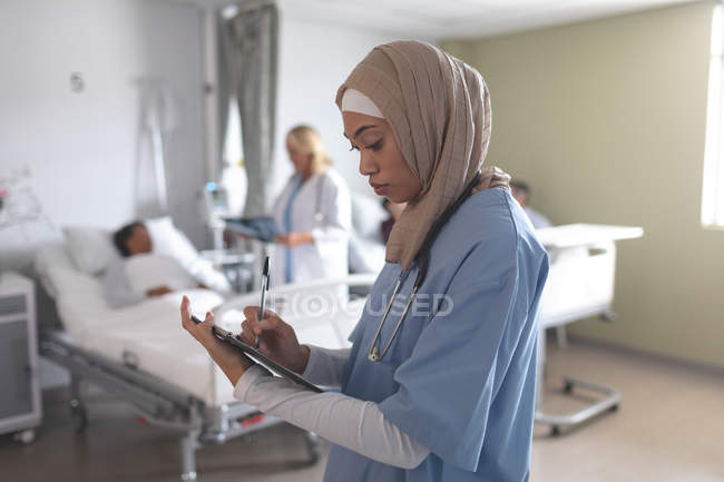 Vista lateral de la doctora de raza mixta en hiyab escribiendo en portapapeles en la sala del hospital. En el fondo diversos médicos están interactuando con sus pacientes . - foto de stock