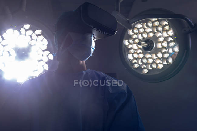 Низький кут зору Кавказького хірурга використанням гарнітури віртуальної реальності в операційній кімнаті в лікарні. Медичні вогні на задньому плані. — стокове фото