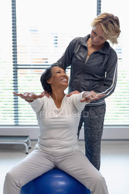 Vue de face du physiothérapeute féminine caucasienne aidant heureuse patiente métisse sur le ballon d'exercice à l'hôpital — Photo de stock