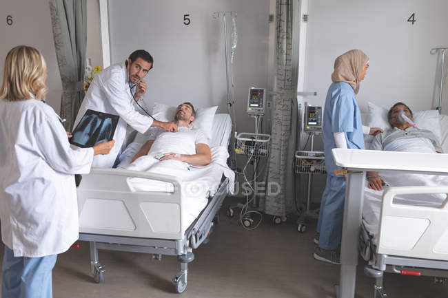 Visão lateral do médico masculino caucasiano examinando o paciente masculino com estetoscópio, enquanto o médico feminino olha para o raio-x na enfermaria no hospital . — Fotografia de Stock