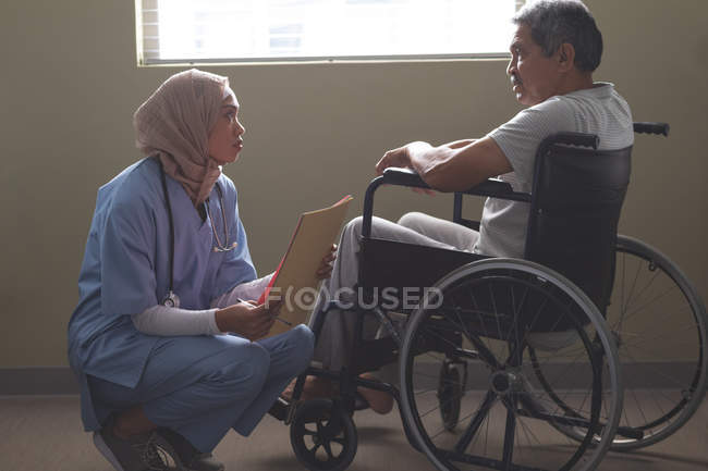 Vista laterale della paziente di razza mista che interagisce con il paziente anziano di razza mista disabile in sedia a rotelle nel reparto ospedaliero . — Foto stock