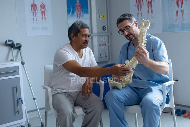 Vista frontale del bel chirurgo ortopedico maschio caucasico che spiega il modello della colonna vertebrale al paziente maschio di razza mista in ospedale — Foto stock