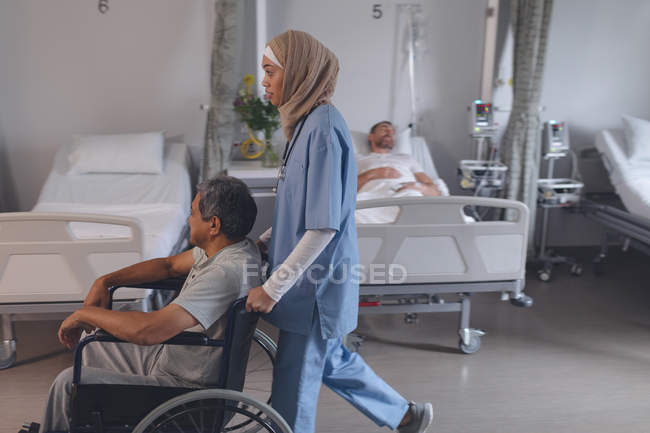 Vista lateral del médico de raza mixta en hijab empujando a un paciente masculino de raza mixta sénior en silla de ruedas en el hospital . - foto de stock