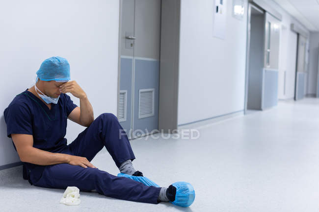 Вигляд на передню частину Кавказького чоловічого хірурга з ручним обличчям сидить у коридорі в лікарні. Хірург носить хірургічну маску, сукню, рукавички і шапочка. — стокове фото