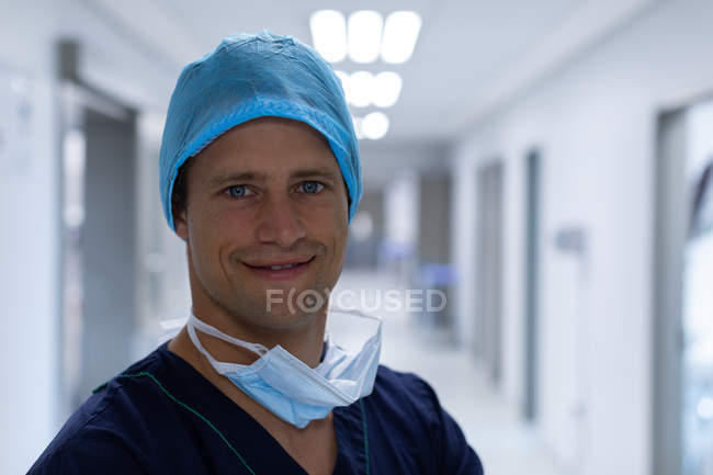 Retrato de um belo cirurgião caucasiano, vestido cirúrgico em pé no corredor do hospital. Está a usar boné e máscara cirúrgica. . — Fotografia de Stock