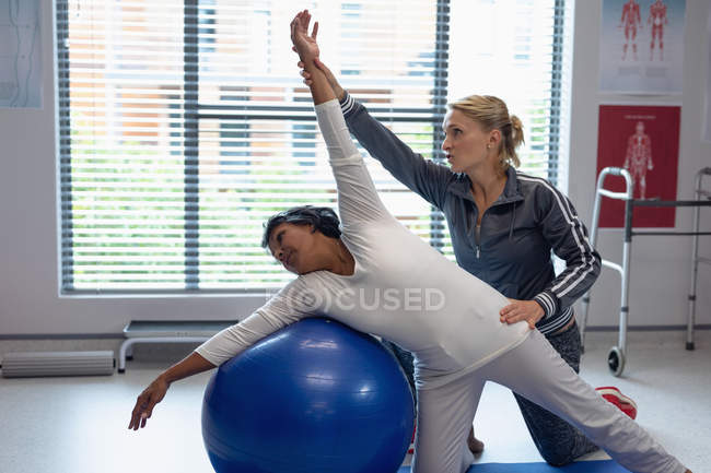 Vue de face du physiothérapeute féminin caucasien aidant une patiente métisse sur le ballon d'exercice à l'hôpital — Photo de stock