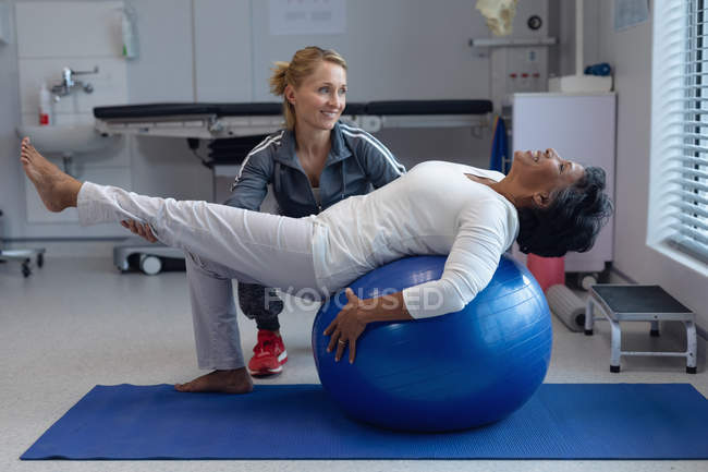 Vista lateral de la fisioterapeuta femenina caucásica feliz ayudando a la paciente de raza mixta en la pelota de ejercicio en el hospital - foto de stock