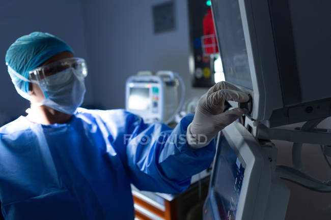 Vista lateral del botón de giro del cirujano femenino de raza mixta del monitor quirúrgico en el quirófano del hospital - foto de stock