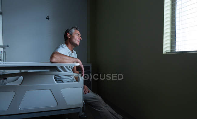 Side view of thoughtful Caucasian male patient sitting on bed while looking outside window in the ward in hospital — Stock Photo