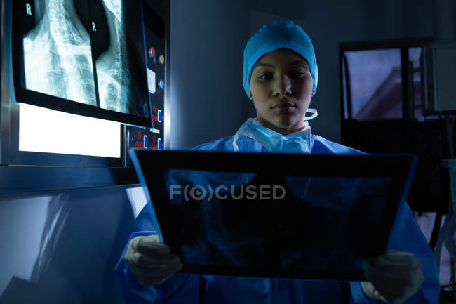 Vista frontal de una joven cirujana de raza mixta examinando rayos X en quirófano en el hospital. El cirujano usa bata quirúrgica, gorra, guantes de látex y máscara. - foto de stock