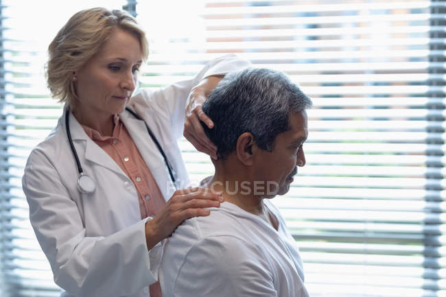 Vista lateral del médico femenino caucásico que examina el cuello de paciente masculino de raza mixta en el hospital - foto de stock