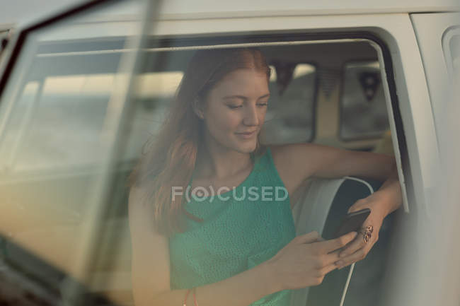 Vista lateral de la hermosa joven mujer caucásica utilizando el teléfono móvil en el asiento delantero de la caravana en la playa - foto de stock