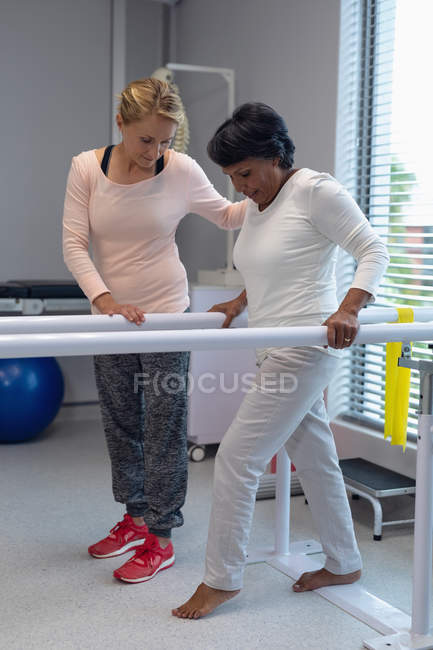 Visão lateral da fisioterapeuta feminina caucasiana ajudando a paciente mestiça a caminhar com barras paralelas no hospital — Fotografia de Stock