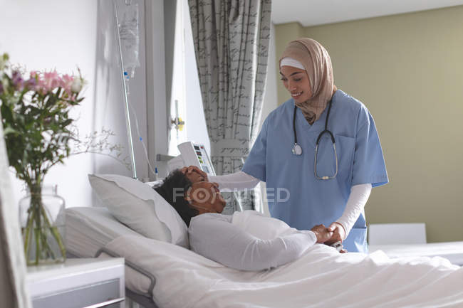 Vista frontal de la hermosa doctora de raza mixta en hijab comprobando la temperatura del paciente femenino de raza mixta con la mano en la sala en el hospital . - foto de stock