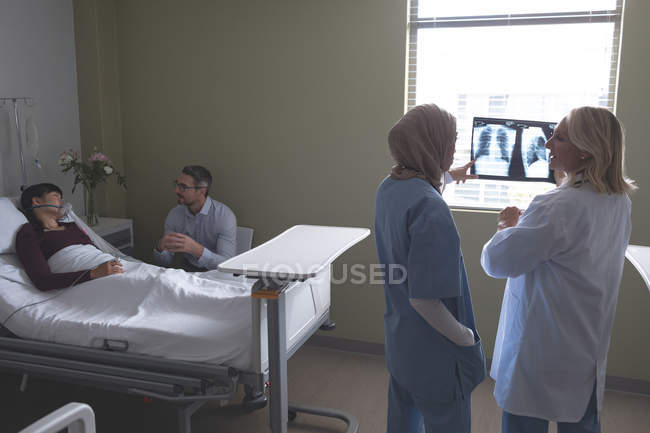 Vista posteriore di diverse medici donne che discutono sopra i raggi X rapporto nel reparto in ospedale. Sullo sfondo asiatica paziente femminile che dorme a letto mentre l'uomo caucasico è seduto accanto a lei nel reparto in ospedale . — Foto stock