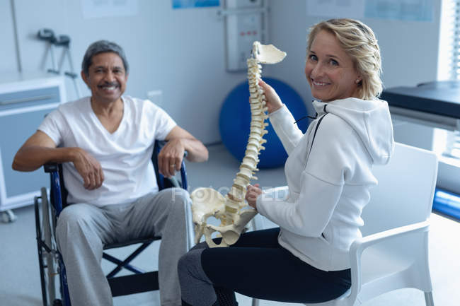 Vue de face du physiothérapeute caucasien expliquant le modèle de colonne vertébrale au patient masculin métis en fauteuil roulant à l'hôpital — Photo de stock