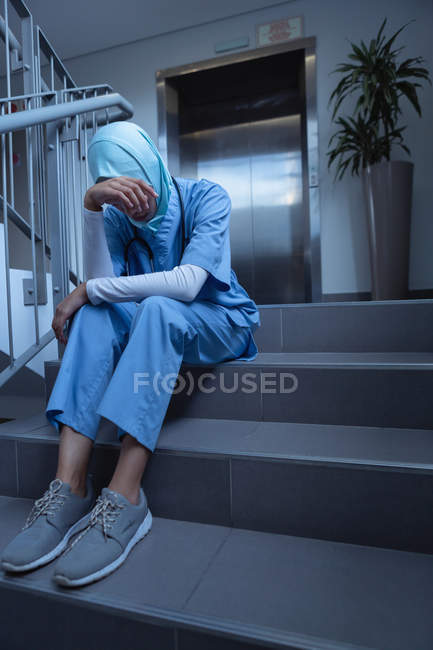 Front view of sad mixed-race female surgeon in hijab with hand on forehead sitting on stairs at hospital — Stock Photo