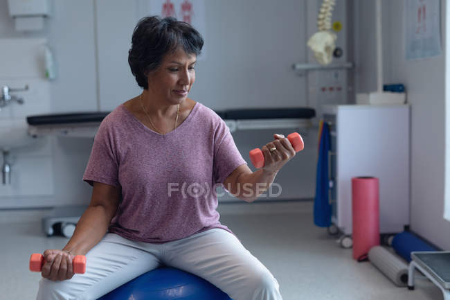 Vista frontal de una hermosa paciente de raza mixta que hace ejercicio con pesas en una pelota de ejercicio en el hospital - foto de stock