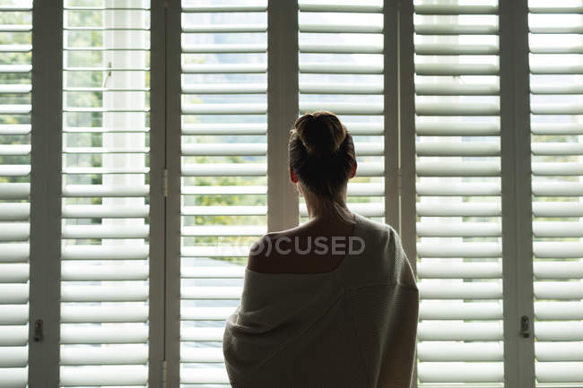 Vista trasera de la mujer caucásica mirando a través de la ventana en casa - foto de stock