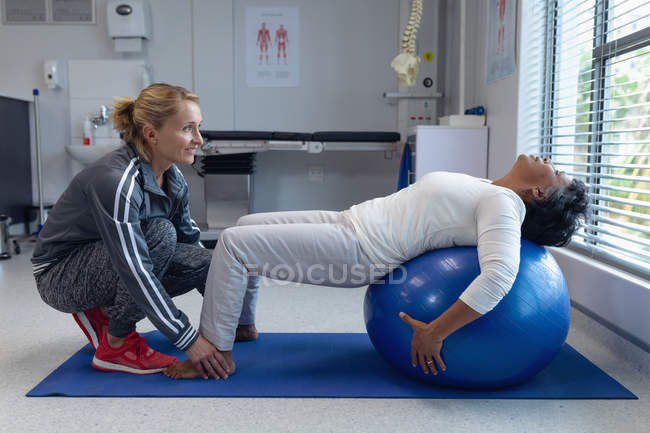 Visão lateral de fisioterapeuta feminina branca dando fisioterapia a paciente mestiça em exercício no hospital — Fotografia de Stock