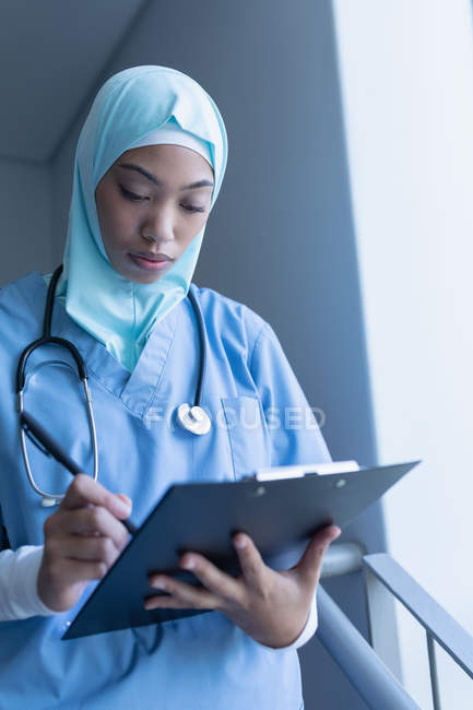 Side view of mixed race female doctor in hijab writing on clipboard at staircase in hospital — Stock Photo