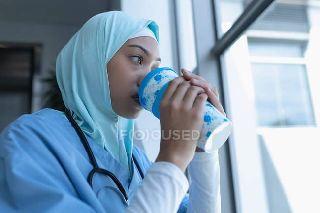 Vista lateral del médico femenino de raza mixta en hijab tomando café en la escalera en el hospital - foto de stock