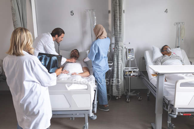 Побочный обзор врача-кавказца, осматривающего пациента-мужчину стетоскопом, в то время как медсестра смешанной расы в хиджабе помогает в палате больницы. Белая женщина-врач проводит рентген и пожилой пациент смешанной расы спит в постели . — стоковое фото