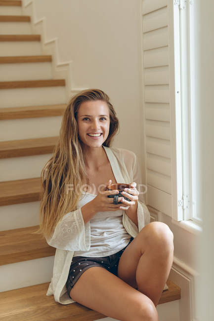 Портрет щасливої кавказької жінки з чашкою кави сидячи на сходах в домашніх умовах — стокове фото