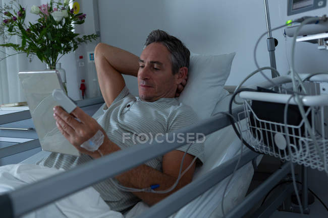 Front view of handsome Caucasian male patient using digital tablet while lying in bed with one hand behind head in the ward in hospital — Stock Photo