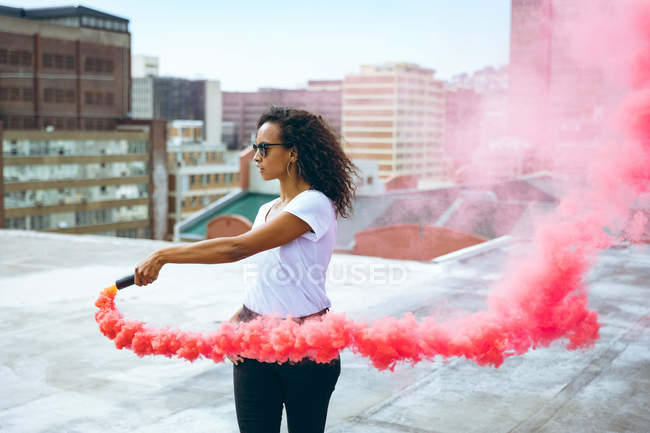 Side view of a young African-American woman wearing a white shirt and eyeglass holding a smoke maker producing red smoke on a rooftop with a view of buildings — Stock Photo