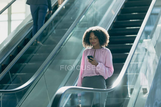 Vista frontal de la mujer de negocios afroamericana mirando el teléfono móvil mientras usa escaleras mecánicas en la oficina moderna - foto de stock
