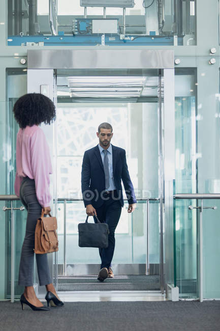 Vista frontal del ejecutivo masculino caucásico saliendo del ascensor en la oficina moderna. Mujer afroamericana esperando para entrar en el ascensor - foto de stock