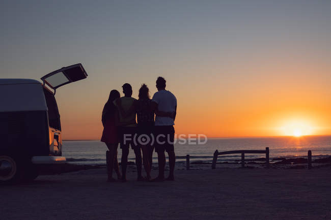 Vista frontal de diversos amigos com braços em torno de pé perto van campista durante o pôr do sol — Fotografia de Stock