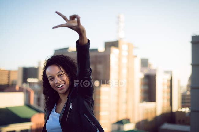 Vista frontal de una joven afroamericana con una chaqueta de cuero mirando a la cámara mientras hace un signo de paz y de pie en una azotea con una vista de los edificios - foto de stock
