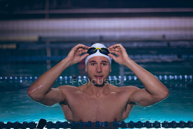 Vue de face d'un homme nageur caucasien tenant les lunettes sur son bonnet de bain blanc alors qu'il se tenait debout dans la piscine — Photo de stock