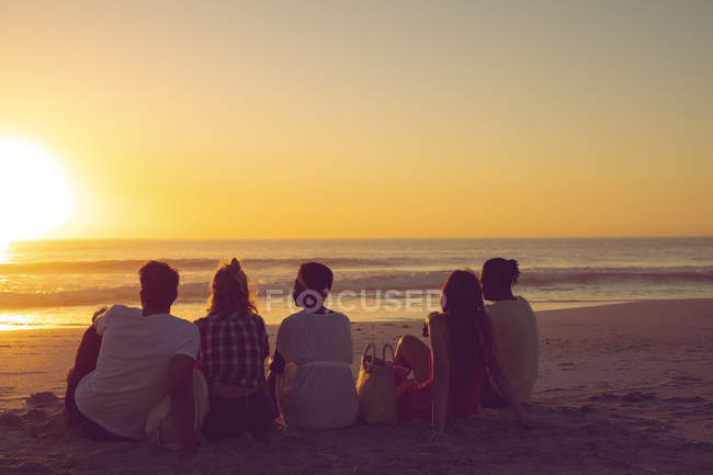 Visão traseira de diversos amigos sentados juntos na praia durante o pôr do sol — Fotografia de Stock