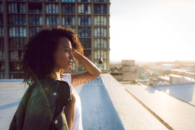 Vista lateral de uma jovem afro-americana com uma jaqueta de couro sobre o ombro olhando para longe da câmera enquanto estava em pé em um telhado com vista para um edifício — Fotografia de Stock