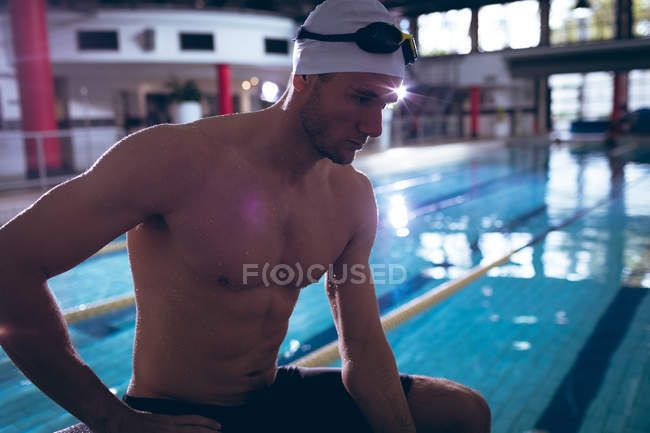 Side view of a male Caucasian swimmer wearing a white swimming cap and goggles sitting by an Olympic sized pool inside a stadium — Stock Photo