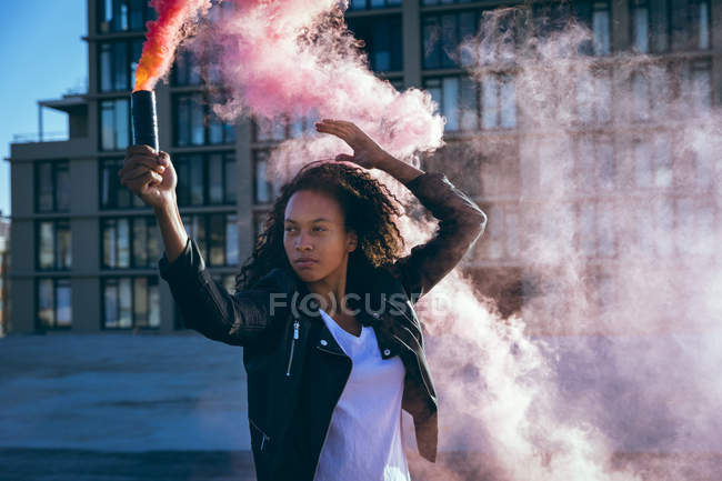 Front view of a young African-American woman wearing a leather jacket holding a smoke maker producing an orange smoke on a rooftop with a view of a building and sunlight — Stock Photo